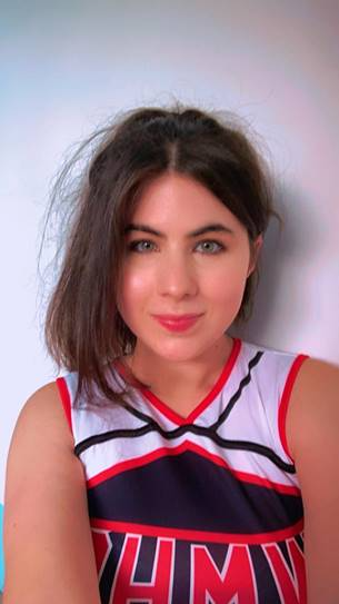 a white woman with green eyes and her straight brown hair tied up in a messy ponytail. She is wearing a cheerleader outfit.